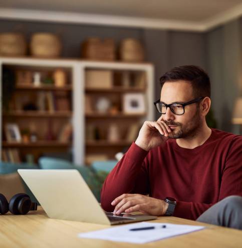 A busy, focused young adult male freelancer sitting at a desk and resting his head on his hand while working on a project on his laptop.